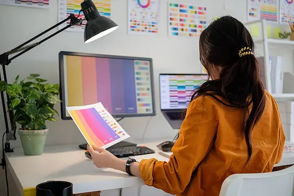 Diseñadora gráfica comparando paleta de colores impresa con el monitor para configurar archivos de impresión